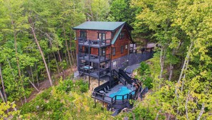A multi-story wooden cabin with a deck and a swimming pool, surrounded by dense forest.