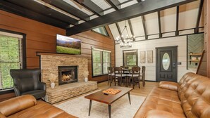 A spacious living room with leather sofas, a stone fireplace, a dining area, and a large TV mounted on the wall.