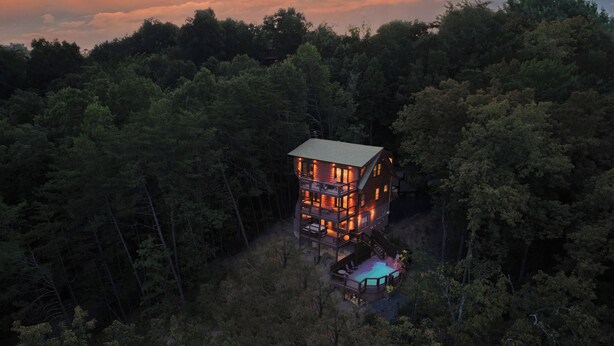 A multi-story wooden cabin surrounded by dense forest, featuring a deck and outdoor seating area.
