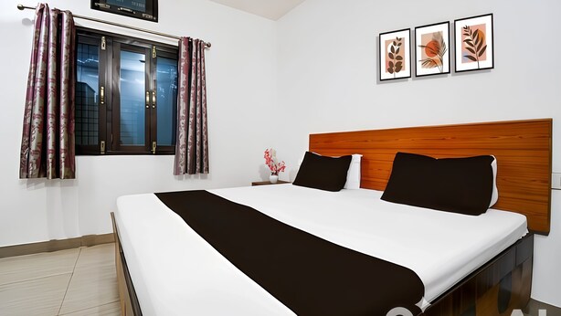 Ein modernes Hotelzimmer mit einem großen Bett, einem hölzernen Kopfteil und einem Fenster mit Vorhängen. An der Wand hängen drei gerahmte Bilder.