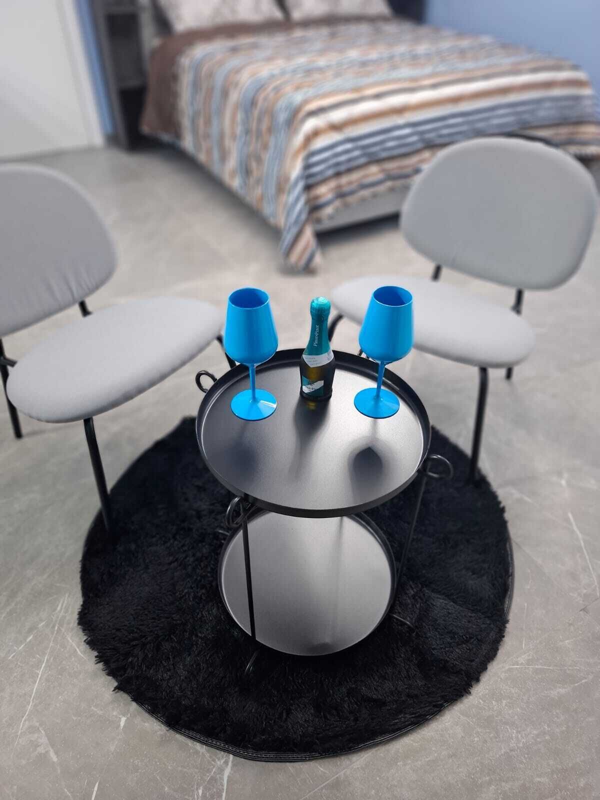 A small round table with three blue cups and a bottle of champagne in the center, surrounded by two grey chairs.