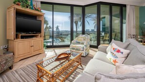 Un salon doté d’un grand téléviseur à écran plat, d’une table basse en osier, d’un canapé et d’un fauteuil. Un balcon, accessible par une porte vitrée, donne sur un espace piscine.