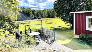 A wooden deck with a table and chairs, a red building with a window, and a fenced green field.