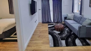 Ein moderner, gut beleuchteter Raum mit einem grauen Sofa, einem gemusterten Teppich und einem Bett mit weißem Kopfteil.