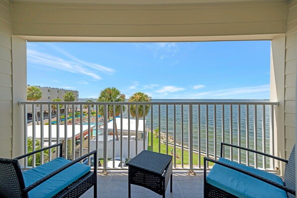 A balcony with two wicker chairs and a small table, overlooking a body of water and a resort area.