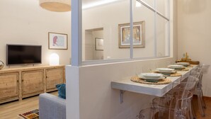 Un salon moderne comprenant un téléviseur, des meubles en bois, un canapé et un coin repas avec des chaises transparentes et une longue table.