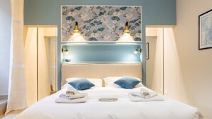 Une chambre d’hôtel avec un lit, deux lampes de chevet, une tête de lit recouverte d’un papier peint à motifs floraux et une fenêtre avec des rideaux.