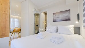 A modern hotel room with a neatly made bed, a dining area with a wooden table and chairs, and a bathroom visible through an open door.