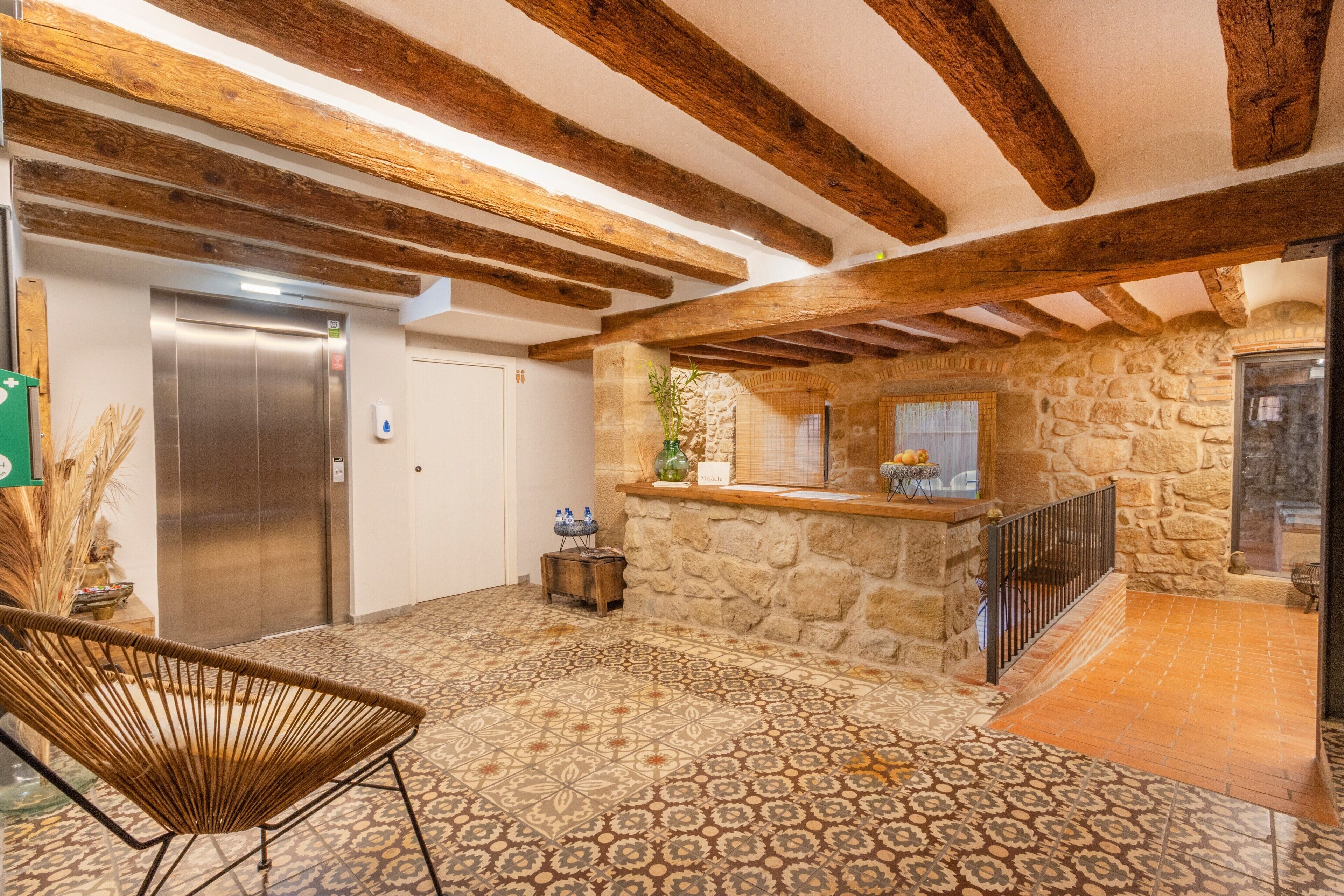 Un vestíbulo con pared de piedra, vigas de madera, un ascensor, una silla de mimbre y suelo estampado.