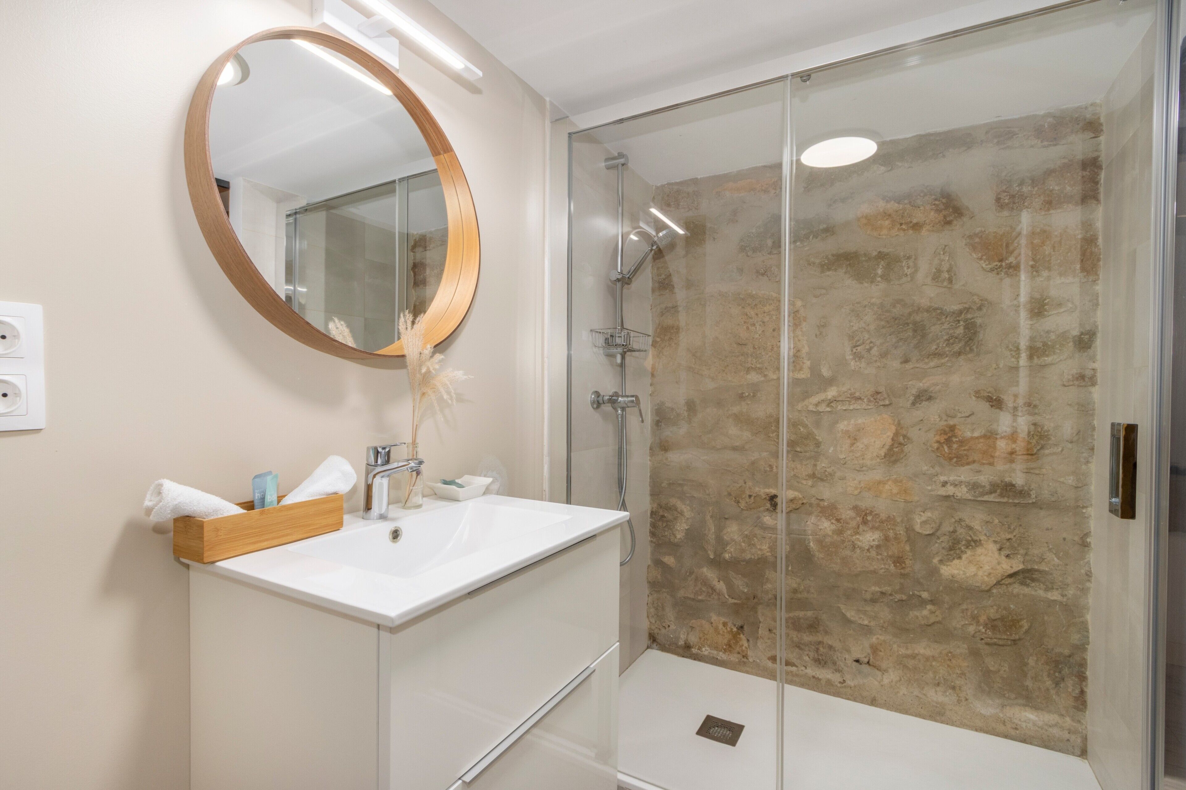 Un baño con lavamanos blanco, tocador de madera, espejo redondo y cabina de ducha con pared de piedra.