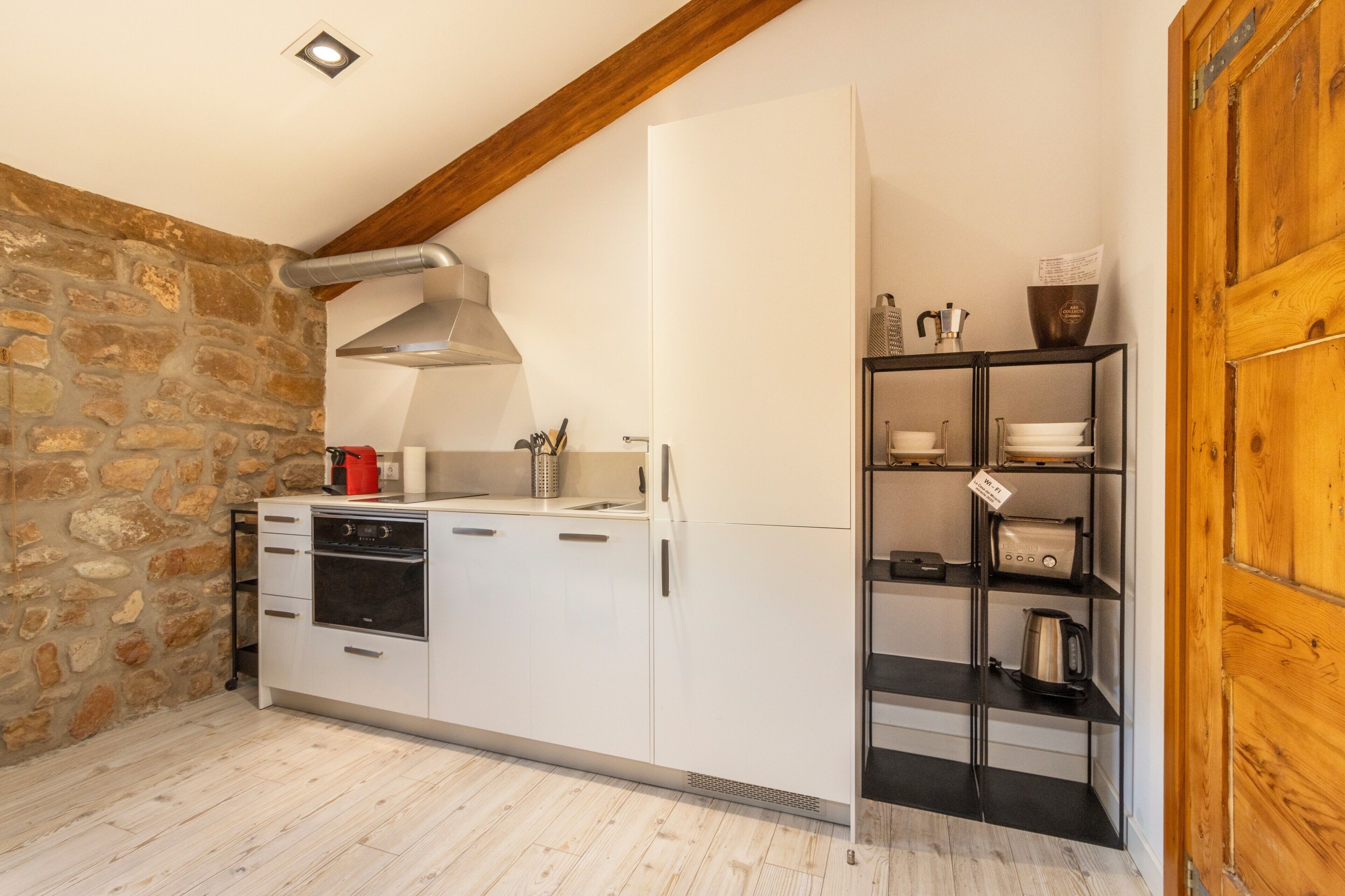 Une cuisine avec des armoires blanches, un four encastré, une hotte de cuisinière en acier inoxydable et une poutre en bois au plafond.