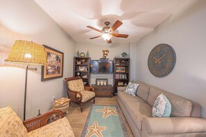 Un salon avec un ventilateur de plafond, un téléviseur, une horloge, un canapé, un fauteuil et un tapis à motifs.