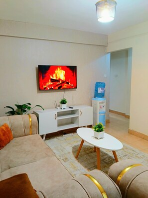 A living room with a flat-screen TV displaying a fire, a white cabinet, a round coffee table, and a beige sofa with gold accents.