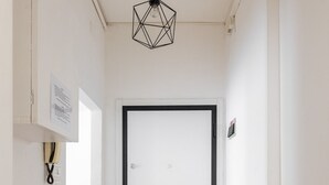 Un pasillo con una puerta blanca, una lámpara colgante de diseño geométrico y un pequeño banco.