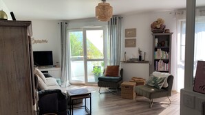 Un salon chaleureux avec un canapé, un fauteuil et une petite table. On y trouve une bibliothèque remplie de livres et d’objets décoratifs. Un téléviseur est fixé au mur, et une fenêtre laisse entrer la lumière naturelle.