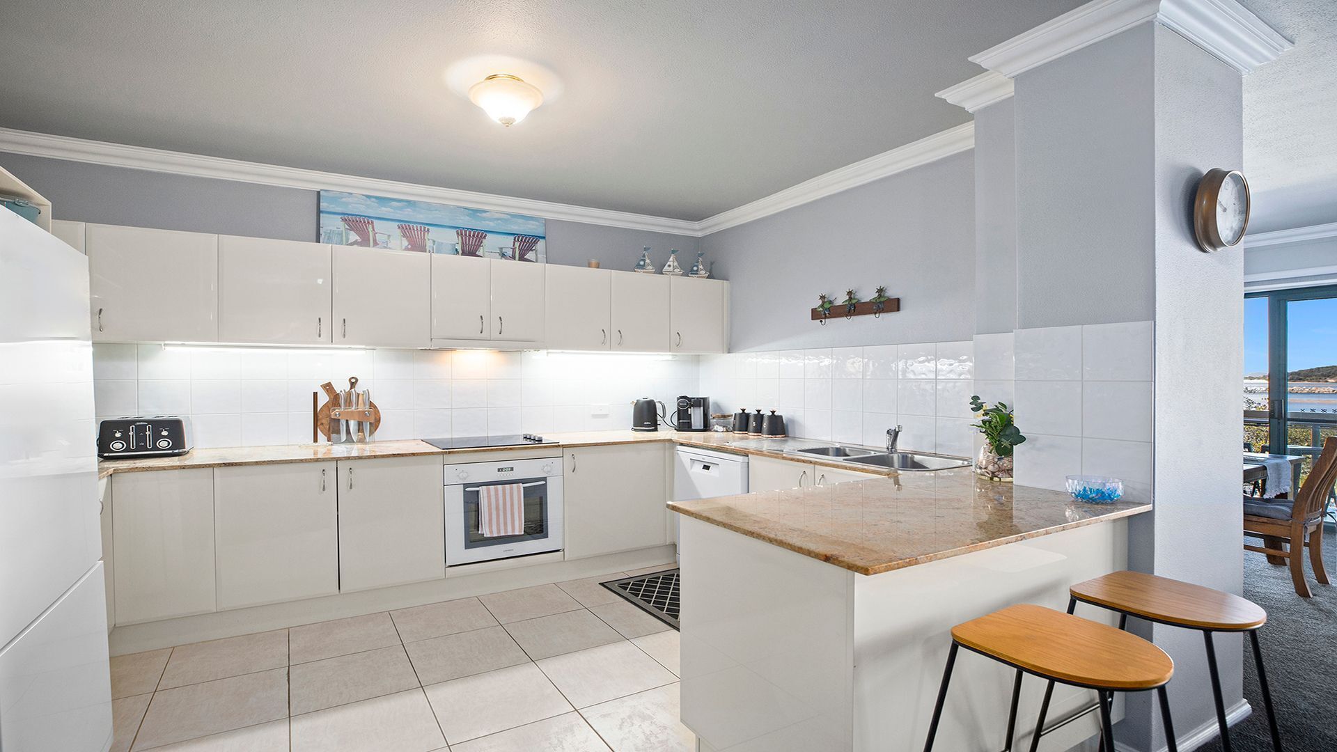 A modern kitchen with white cabinetry, a central island with stools, a built-in oven, and a view of the outdoors through a window.