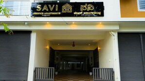 Entrada de un edificio con piso a cuadros, un cartel con 'SAVI SERVICE APARTMENTS' e información de contacto, y una cámara de seguridad.