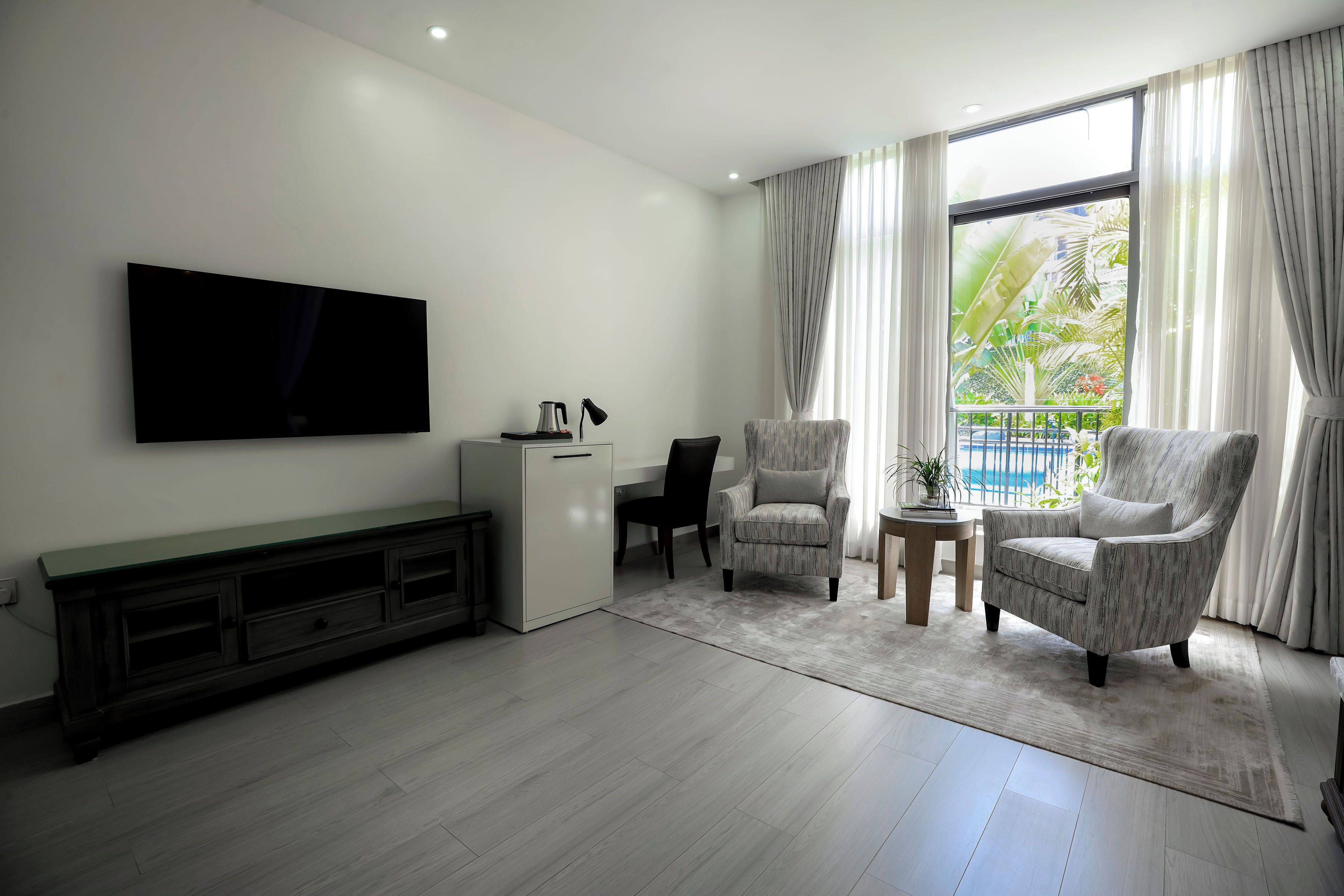 Una sala de estar moderna con un televisor de pantalla plana, un escritorio pequeño, dos sillones, una mesa de centro y un ventanal con cortinas.