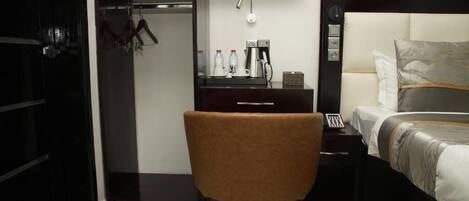 Habitación de hotel con cama, escritorio con cafetera, silla, armario y un cuadro en la pared.