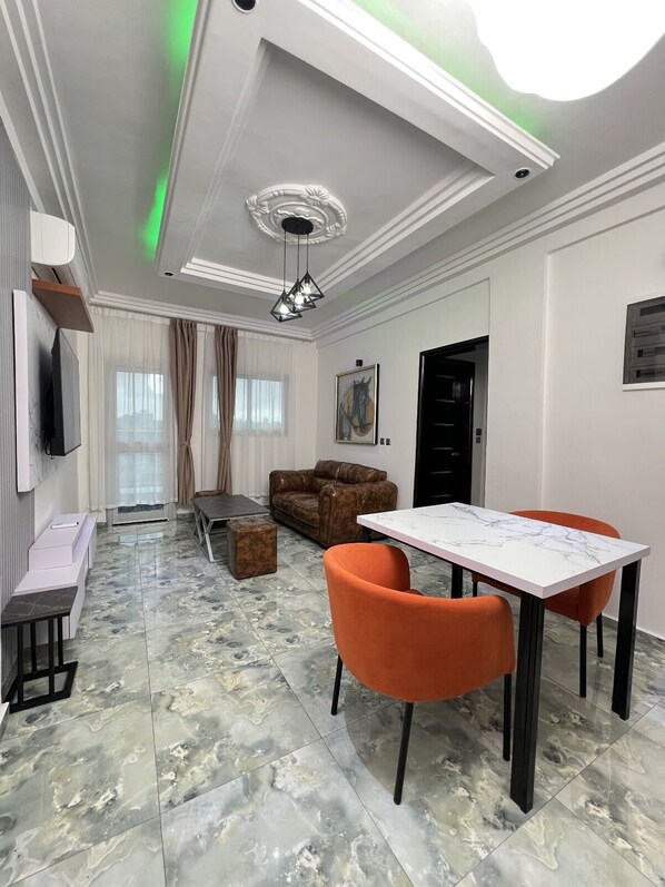 A modern living room with a marble floor, a dining table with orange chairs, a leather sofa, and a TV mounted on the wall. - Exotic Plaza Hotel (Douala)
