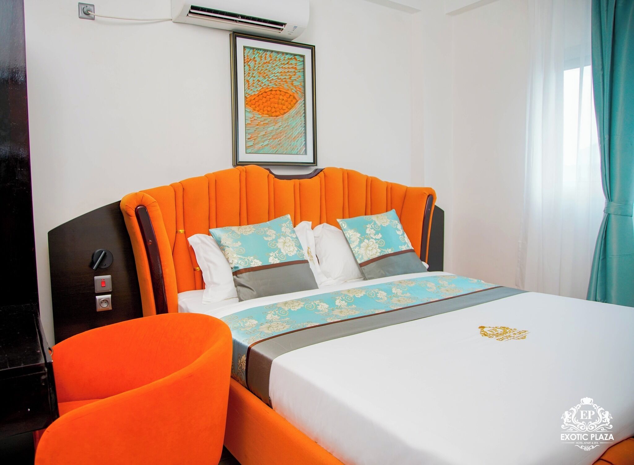 A hotel room with a bed, an orange upholstered chair, a framed artwork on the wall, and a window with curtains.