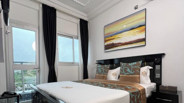 A modern bedroom with a large bed, a nightstand, a bedside table, a painting, and a view of the outdoors. - Exotic Plaza Hotel (Douala)
