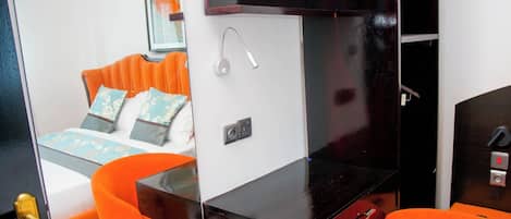 Una habitación de hotel compacta con una cama, una mesita pequeña y dos sillas color naranja.
