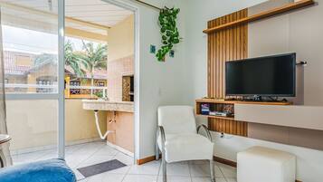 Ein Wohnzimmer mit Fernseher, einem weißen Sessel, einer Couch und einem Balkon mit Blick auf Palmen.