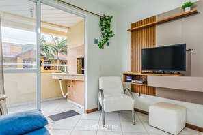 Een woonkamer met een televisie, een witte stoel, een bank en een balkon met uitzicht op palmbomen.