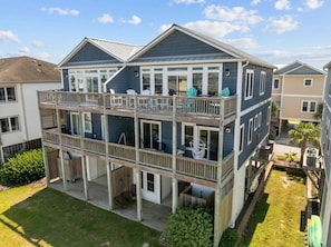 A two-story residential building with a balcony on the upper floor, a covered porch on the lower floor, and a small yard with a palm tree. - You're in Heaven #198 - Boat Dock, Pool, Hot Tub, Dog Friendly, Sleeps 9! (Topsail Beach)