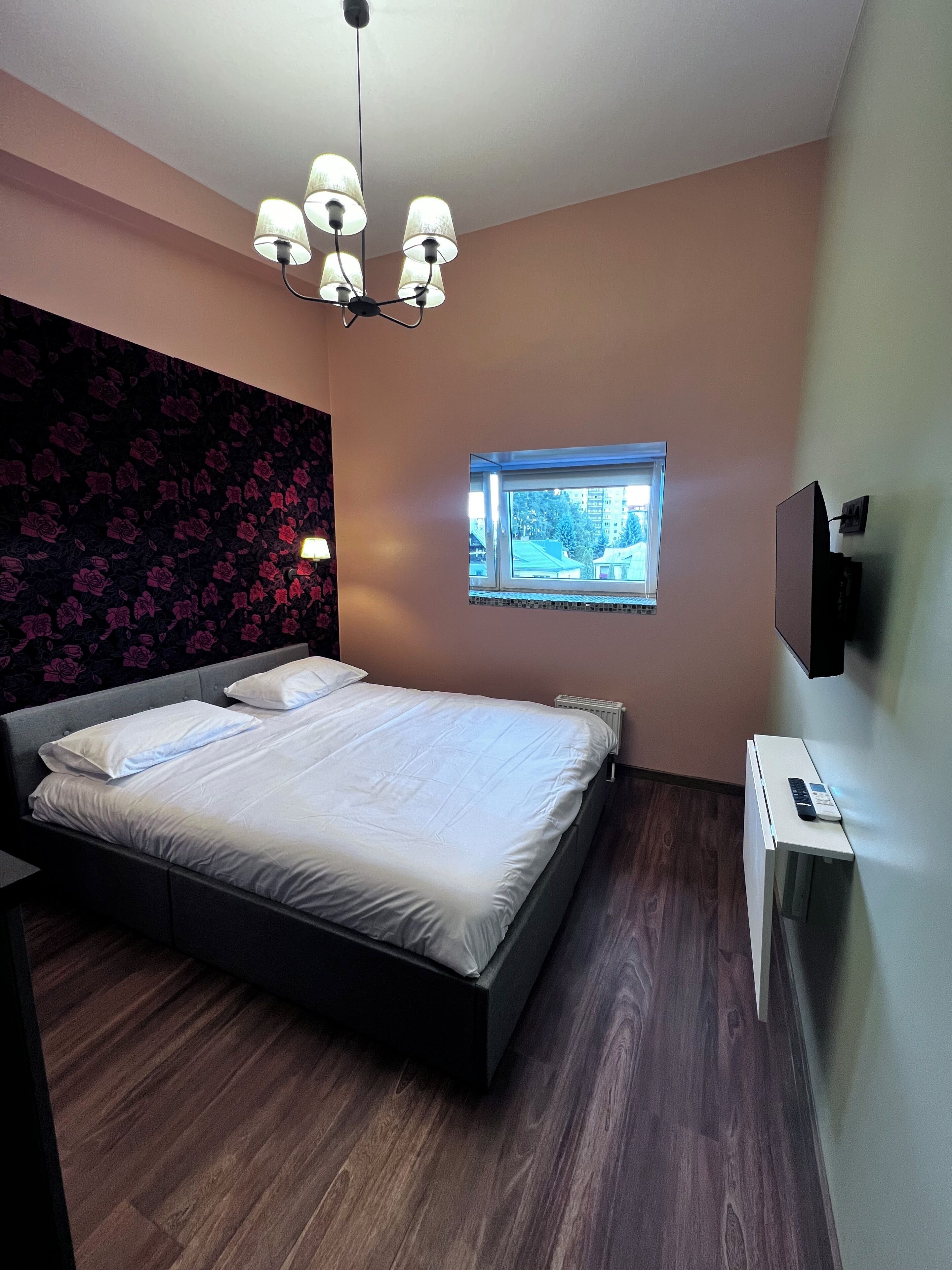 Un dormitorio con cama, ventana, televisor de pared y lámpara en el techo.