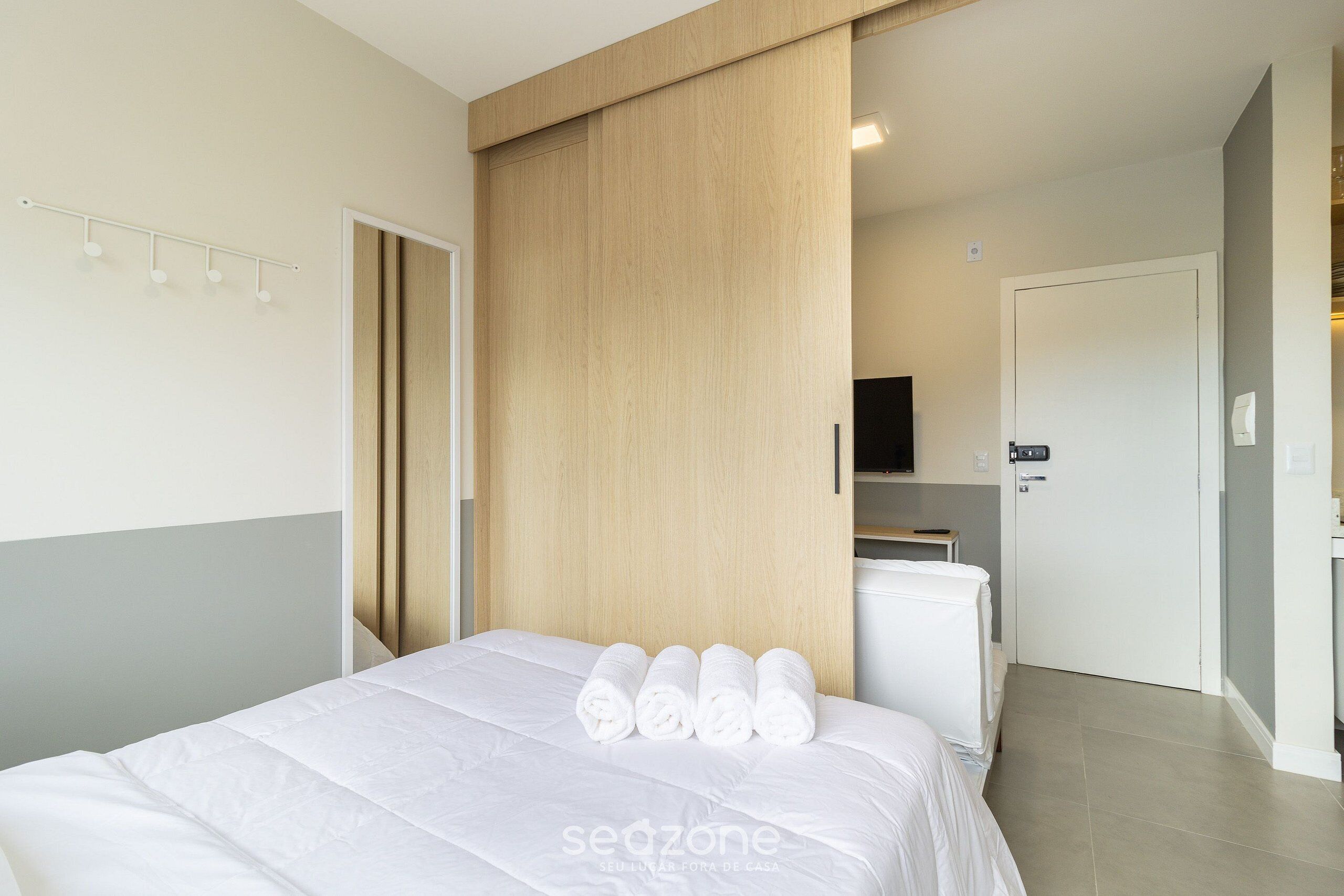 A modern hotel room with a bed, a wooden wardrobe, a flat-screen TV, and a small kitchenette.