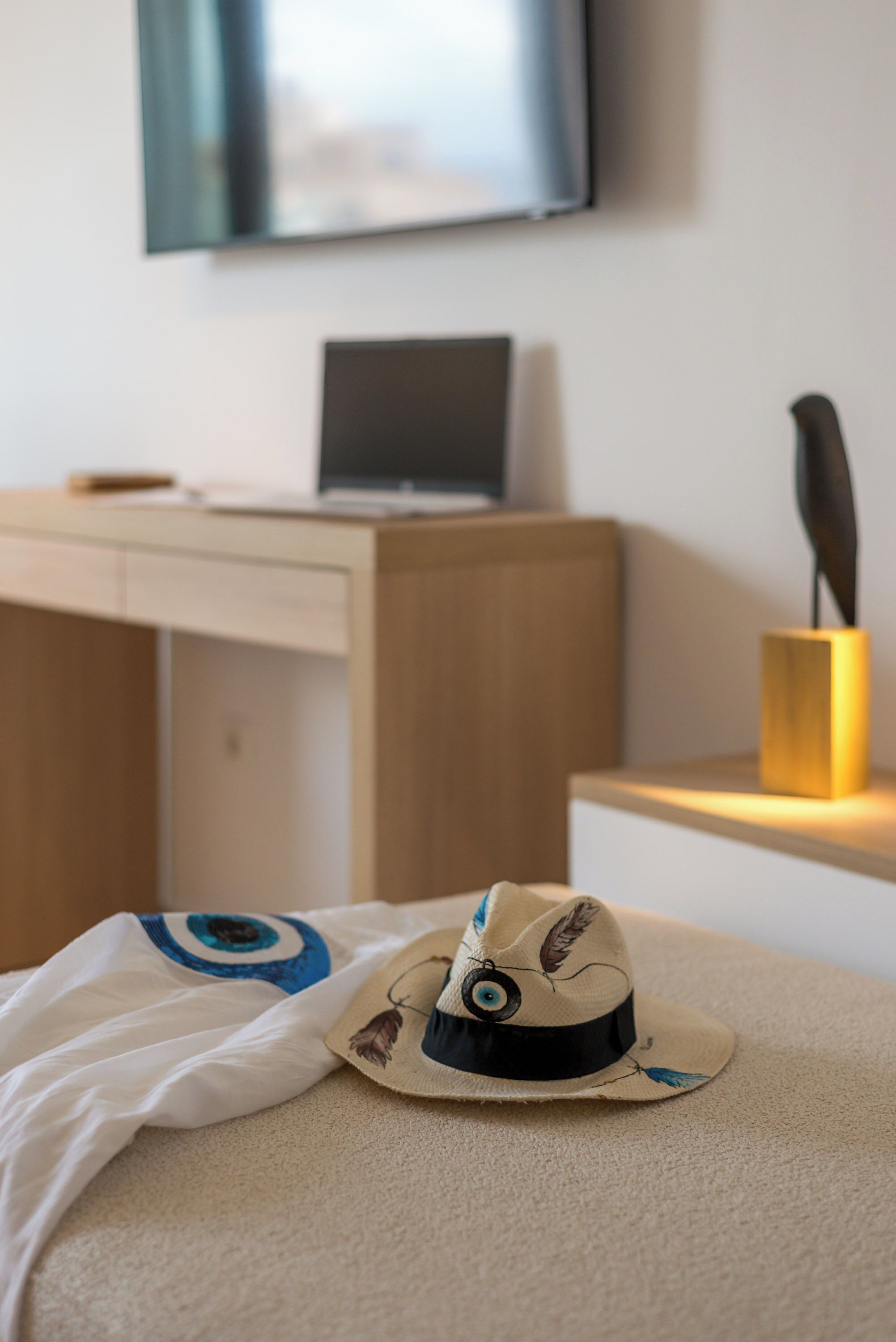 Una cama con un sombrero y una almohada, un escritorio con una computadora portátil y un televisor en la pared.