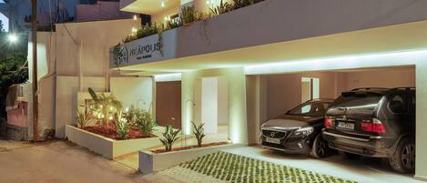 A modern building with a sign reading "NEAPOLIS". It features two cars parked in a covered area, a landscaped garden, and a flat roof with visible structural beams.