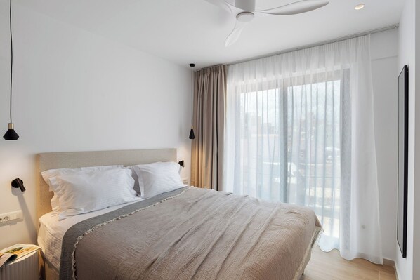 Ein modernes Hotelzimmer mit einem großen Bett, einem Deckenventilator, Blick auf die Stadt durch transparente Vorhänge und einem an der Wand befestigten Fernseher.