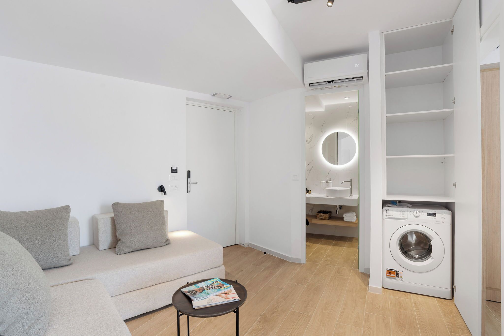 A modern living room with a sofa, a small table with magazines, a washing machine, and a built-in shelving unit.