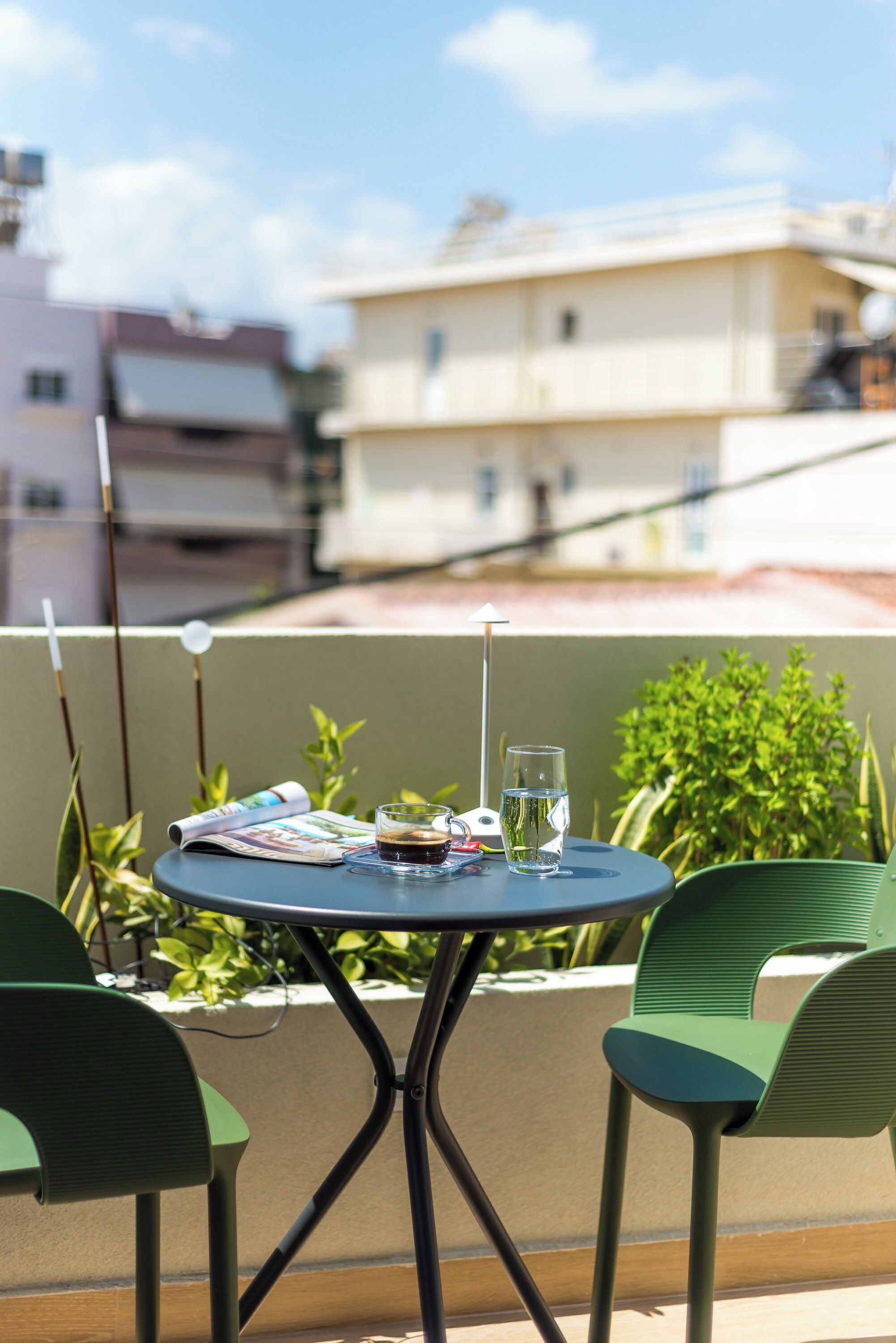 A round table with two green chairs, a glass of water, a magazine, and a bottle of soda on a balcony with a view of buildings and greenery.