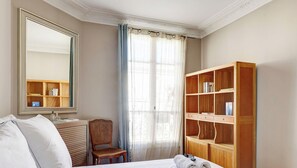 A hotel room with a bed, a wooden chair, a large mirror, a bookshelf, and a window with curtains.