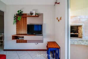 Un salon avec un téléviseur, des étagères en bois, une petite table et un coin cuisine avec un revêtement mural en carreaux.