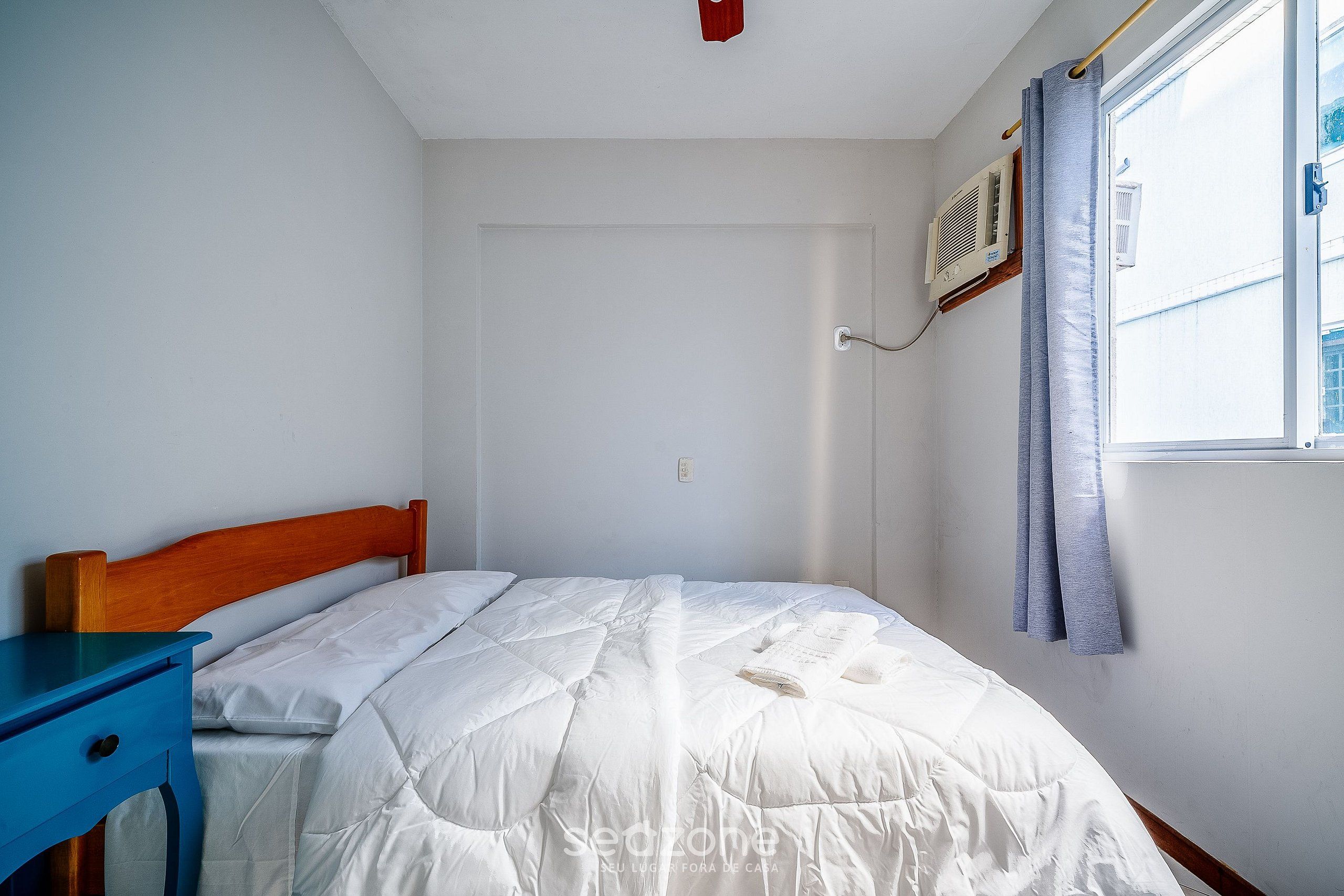 Une chambre à coucher avec une tête de lit en bois, une table de chevet bleue, un lit blanc avec une courtepointe, une fenêtre avec un rideau et un appareil de climatisation fixé au mur.