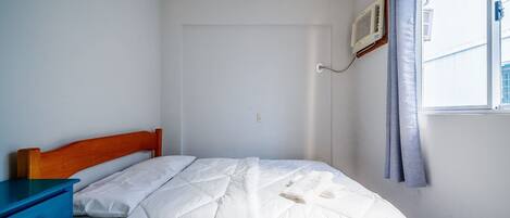 Un dormitorio con cabecera de madera, mesita de noche azul, cama blanca con colcha, ventana con cortina y unidad de aire acondicionado en la pared.