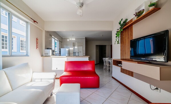 A modern living room with a white sofa, a red accent chair, a TV mounted on a wooden panel, and a kitchen in the background. - Apt W/barbecue 3 Minutes From Bombas Beach Fdl101 (Bombinhas)