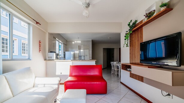 A modern living room with a white sofa, a red accent chair, a TV mounted on a wooden panel, and a kitchen in the background. - Apt W/barbecue 3 Minutes From Bombas Beach Fdl101 (Bombinhas)