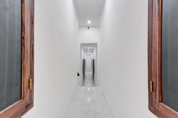 A narrow, well-lit hallway with white walls and a marble floor, leading to a door at the end.