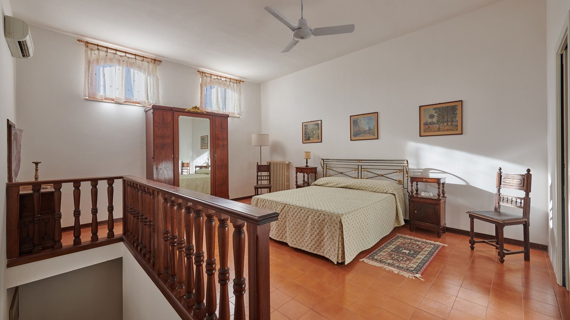 A bedroom with a bed, a chair, a nightstand, a window with curtains, a ceiling fan, and a staircase.