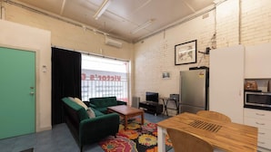 A compact living space with a green sofa, a wooden dining table, a small kitchen area, and a window with blinds.