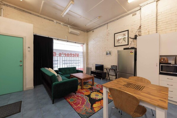A compact living space with a green sofa, a wooden dining table, a small kitchen area, and a window with blinds. - Spacious Studio in Quiet Northcote Neighbourhood (Northcote)