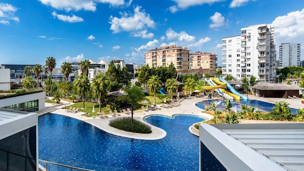 A resort with a large swimming pool, palm trees, and a water slide. - Maxihomes Sirella (Antalya)