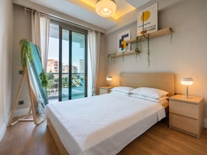 A modern hotel room with a large bed, bedside tables, a view of a cityscape, and a ceiling light fixture. - Maxihomes Sirella (Antalya)