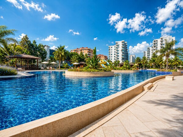A large swimming pool with a clear blue water, surrounded by palm trees and a lounge area with umbrellas. - Maxihomes Sirius Town (Antalya)